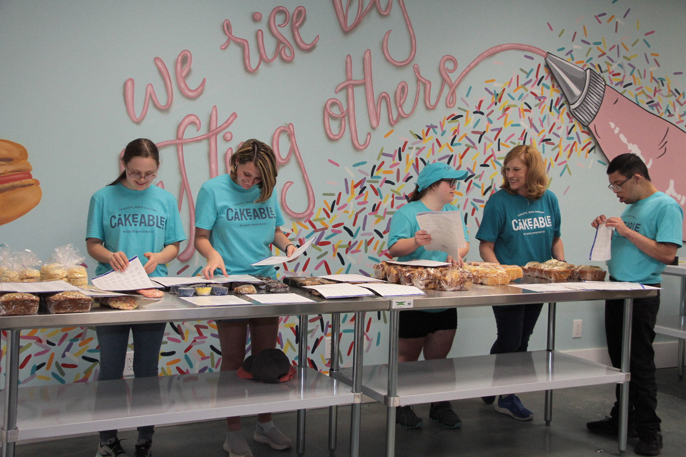 Cakeable team members packaging baked goods along a work table beneath a mural reading "We rise by lifting others"