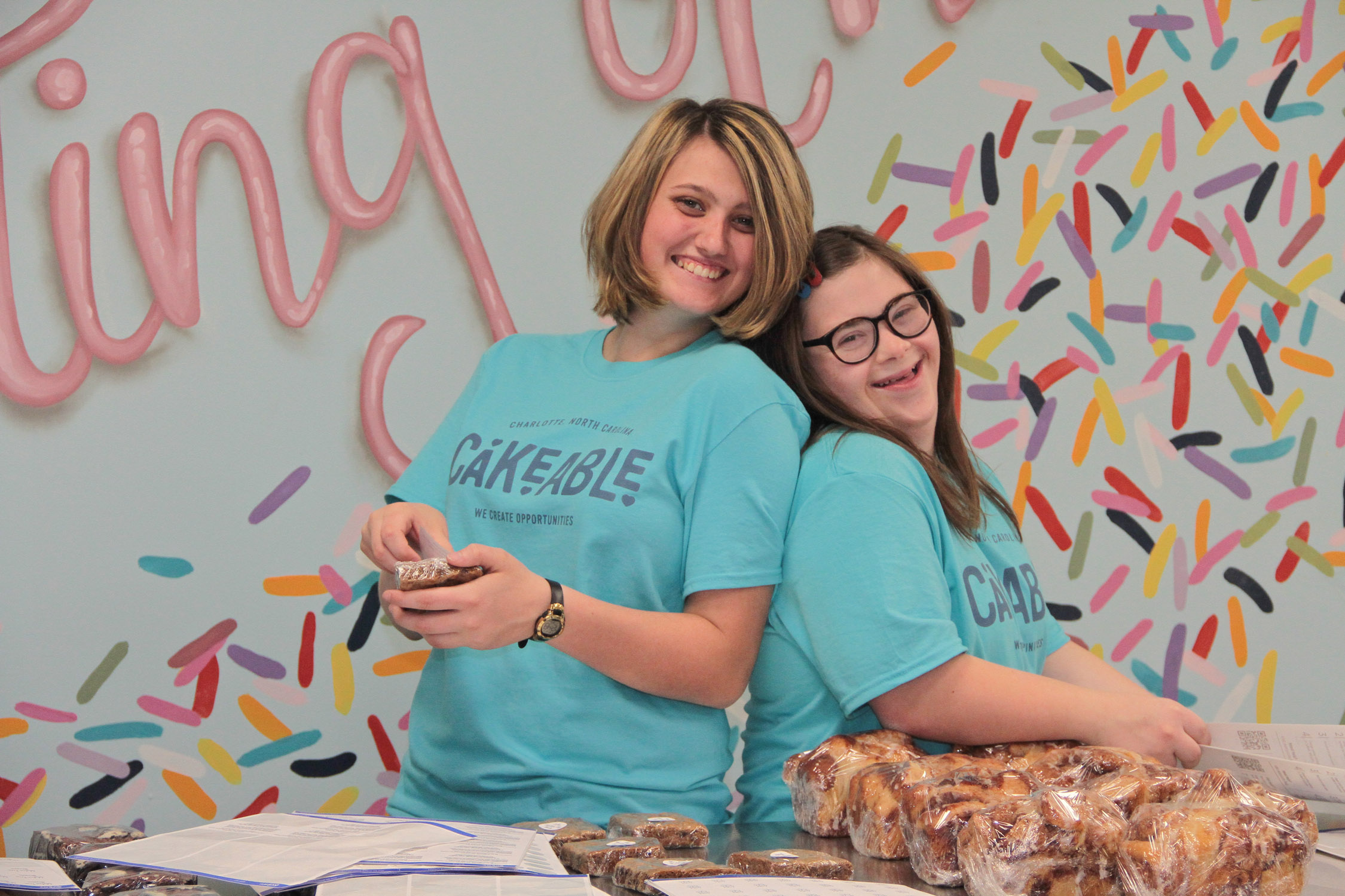 Two Cakeable team members packaging baked goods at a table in front of a sprinkle-patterned wall