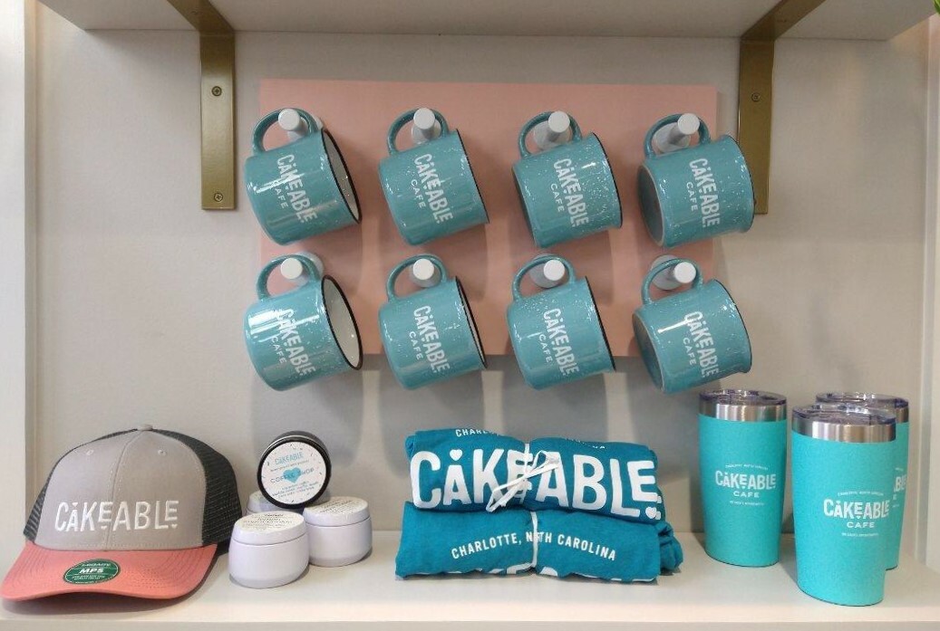 Cakeable Cafe merchandise on a shelf: enamel mugs, a cap, candles, a towel and insulated tumblers