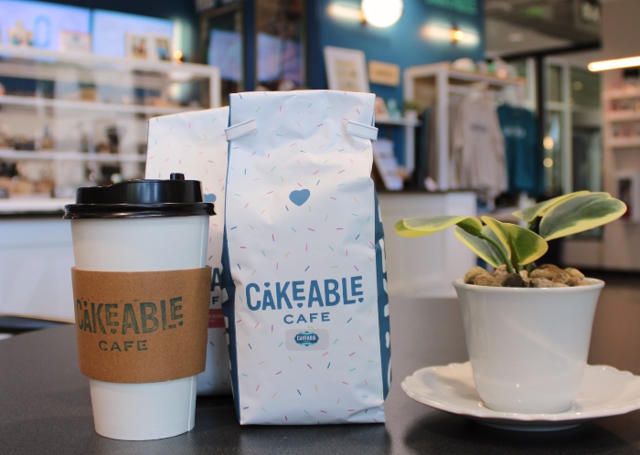 A Cakeable Cafe coffee cup and a bag of Cakeable coffee beans on a counter beside a small potted plant